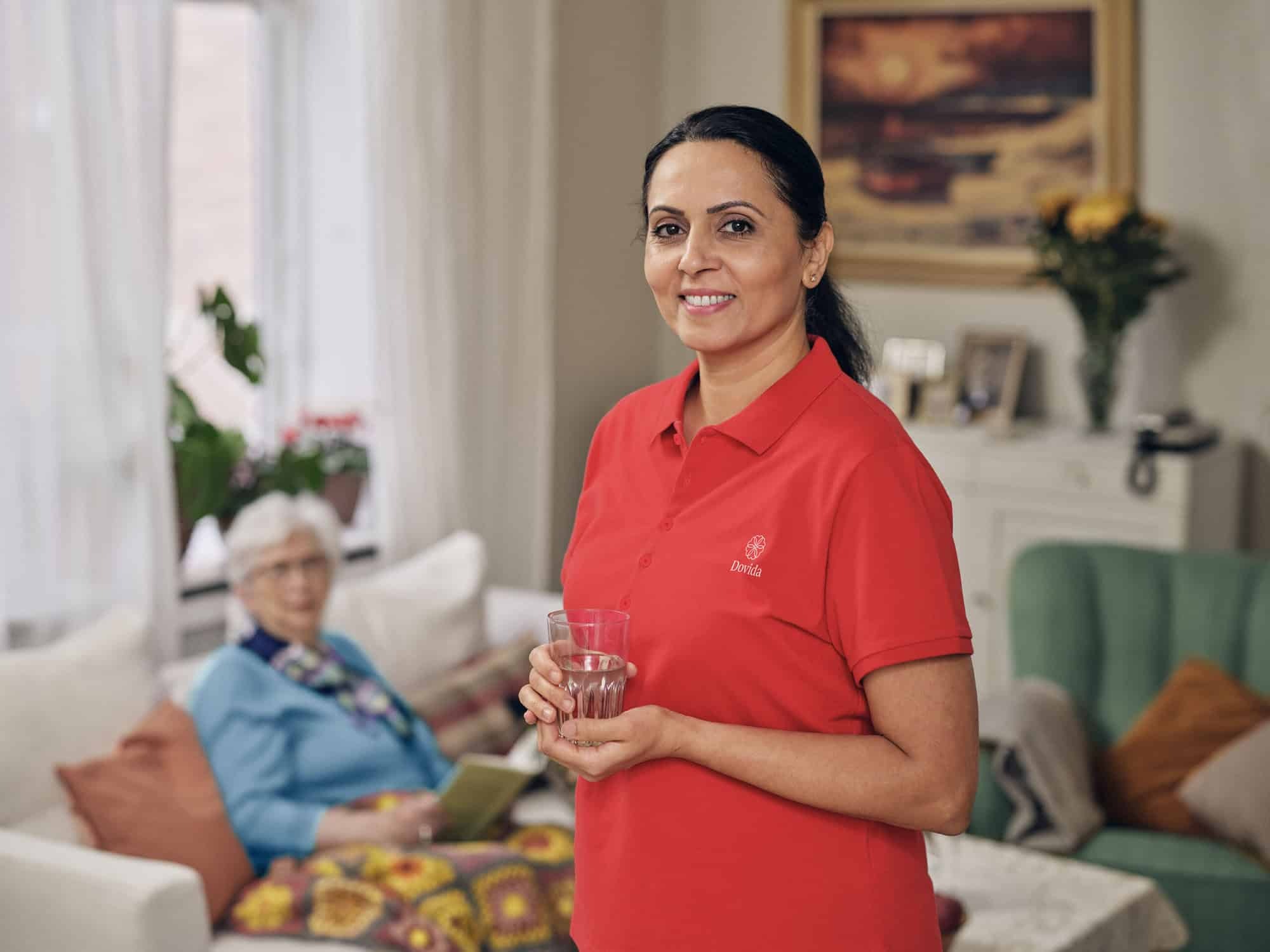 A caregiver dressed in a red Dovida shirt holds a glass, representing the compassionate support offered by personal caregivers at home.