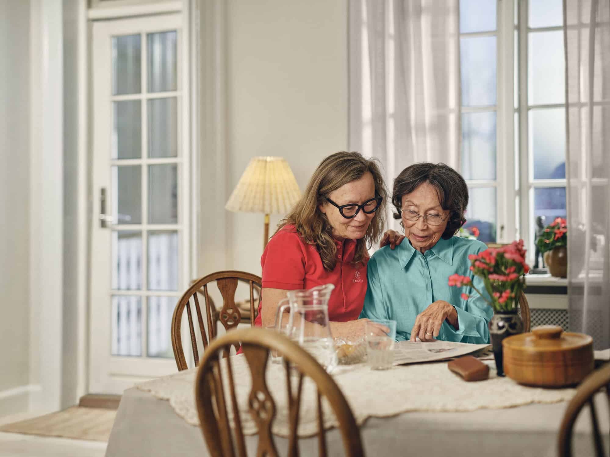 A caregiver and an elderly woman enjoying a conversation at a table, representing companionship and support at home.