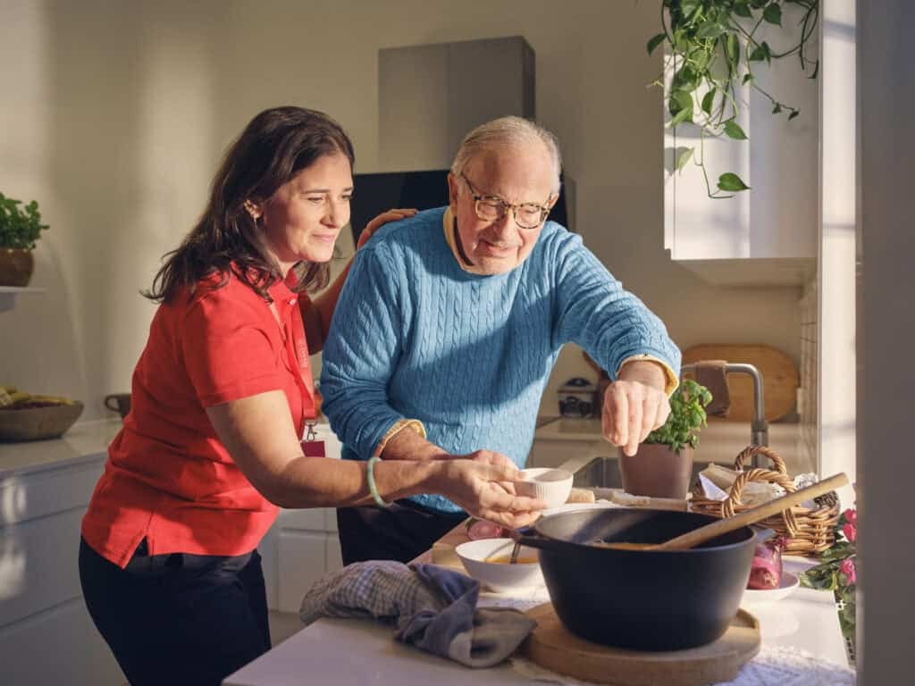 Senior man cooking with support from Dovida Caregiver in a bright kitchen