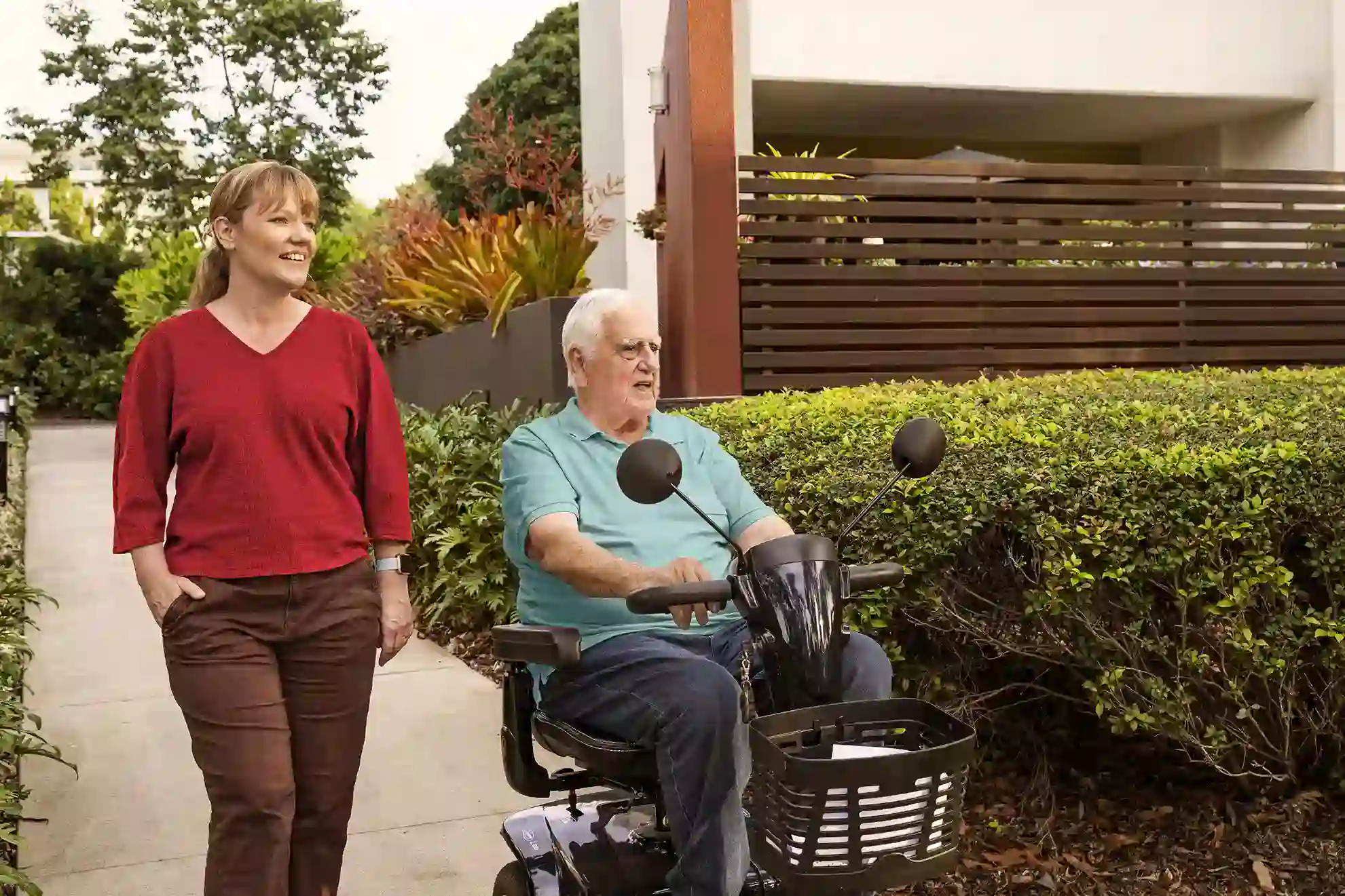 Elderly man driving a mobility scooter outdoors while a caregiver walks beside him along a garden path.